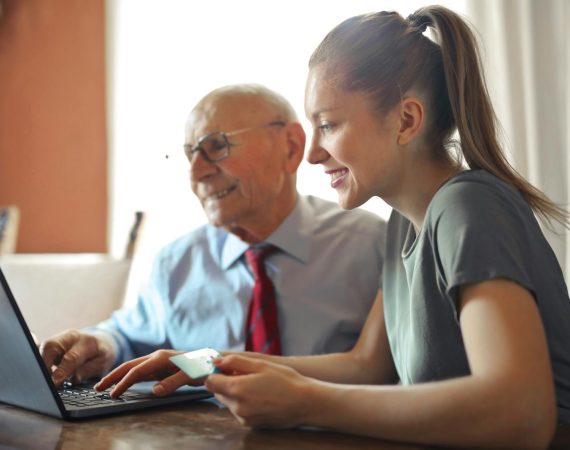 Young woman in casual clothes helping senior man in formal shirt with paying credit card in Internet using laptop while sitting at table