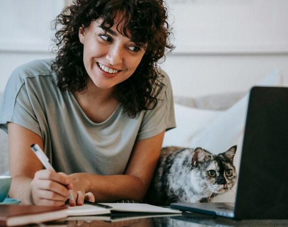 Crop young positive female smiling and taking notes in organizer while cat watching on netbook at table at home