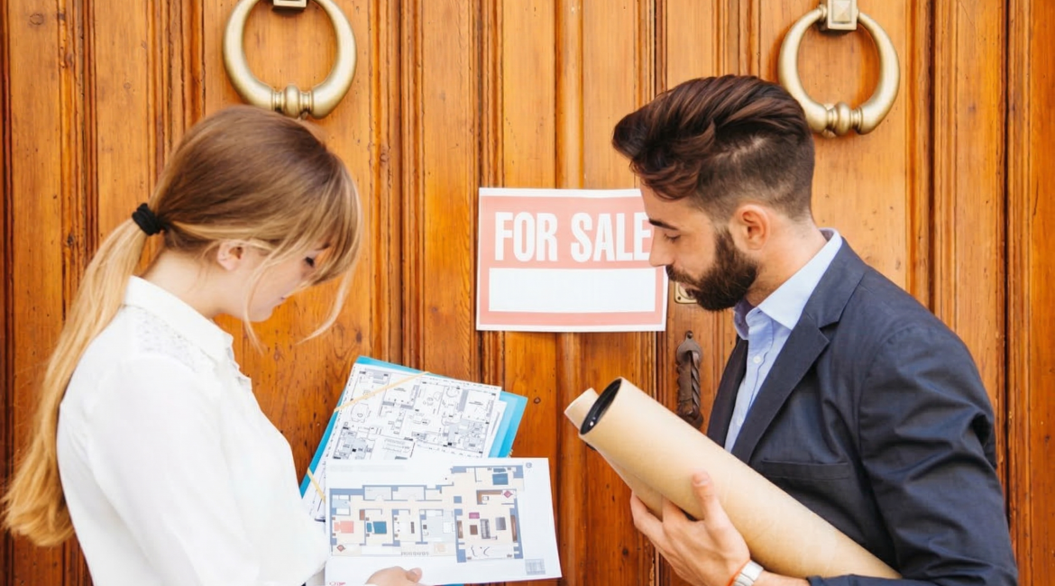 Estate agents reviewing floor plans outside a property with a 'For Sale' sign