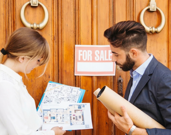 Estate agents reviewing floor plans outside a property with a 'For Sale' sign