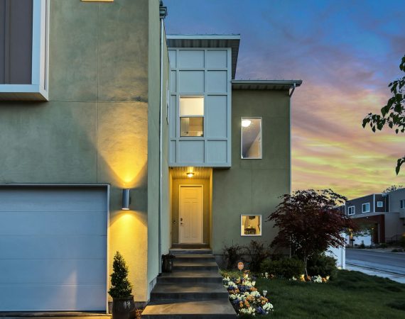 Modern two-storey house exterior with illuminated entrance, manicured garden, and garage at dusk