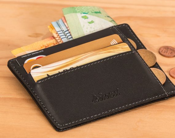 Black leather wallet with cash, credit cards, and coins spread around on a wooden table surface