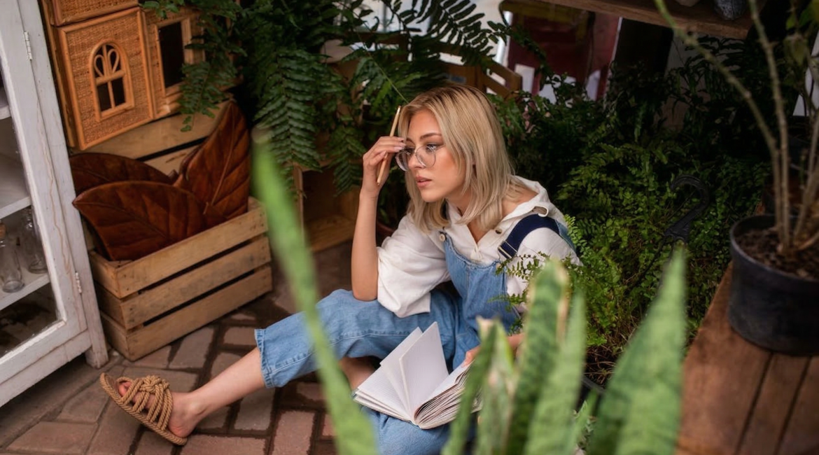 Woman in overalls sitting among plants with a book, adjusting her glasses, in a cosy room setting