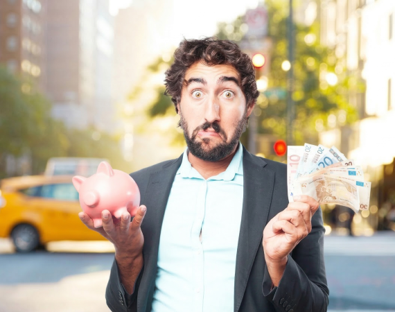 Man holding pink piggy bank and euro notes in city street, looking surprised