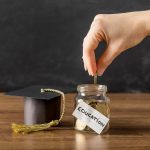 Hand placing coin in education savings jar, graduation cap on table