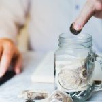 Person adding coins to a jar with banknotes, while using a calculator for budgeting and savings management