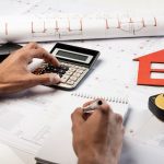 Person calculating construction costs with calculator, notes, and house model on architectural blueprints