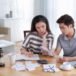 Couple reviewing finances at home, calculating expenses with a notebook, documents, and laptop on desk