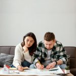 Couple planning home renovation together, reviewing blueprints and taking notes on a sofa with a small model house nearby