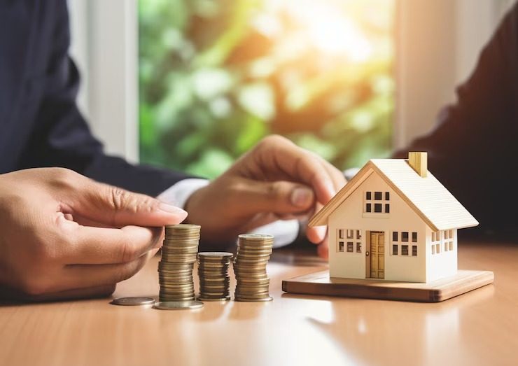 Miniature house next to stacked coins on table, hands in background discussing real estate investment strategy