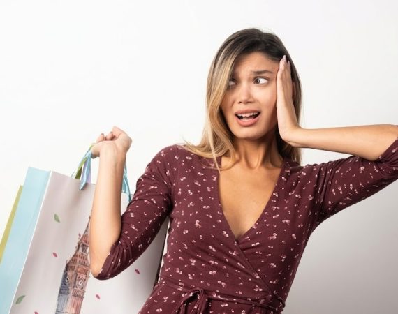 Woman in maroon dress looking surprised, holding colourful shopping bags against a plain white background
