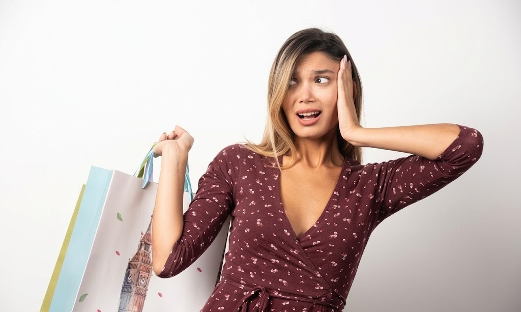 Woman in maroon dress looking surprised, holding colourful shopping bags against a plain white background