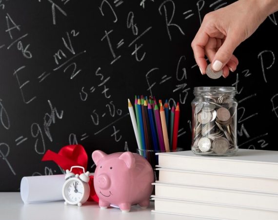 Woman's hand placing coins into a jar beside books and a piggy bank on a blackboard background