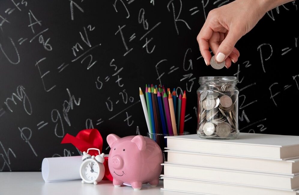 Woman's hand placing coins into a jar beside books and a piggy bank on a blackboard background