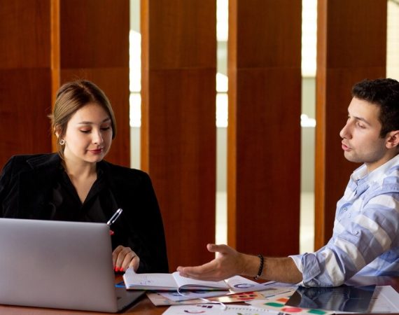 A man and woman sit at a table, focused on a laptop, engaged in discussion or collaboration