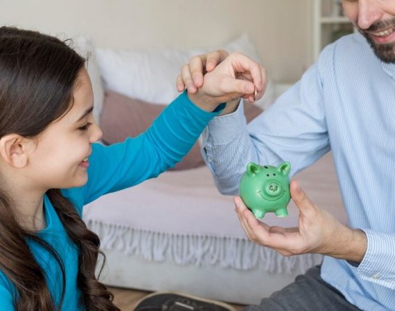 A man and a little girl are smiling as they hold a colorful piggy bank, representing their shared savings goal