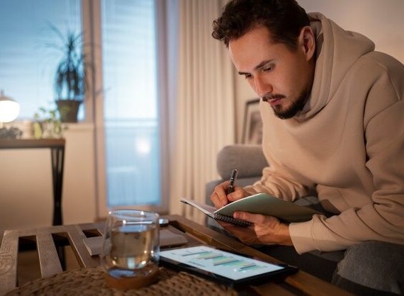 A man sits on a couch, focused on writing in a notebook with a pen in hand
