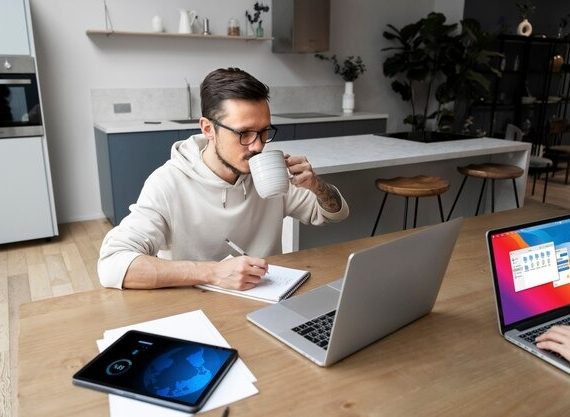 Two individuals focused on their laptops at a table, with a kitchen visible in the background
